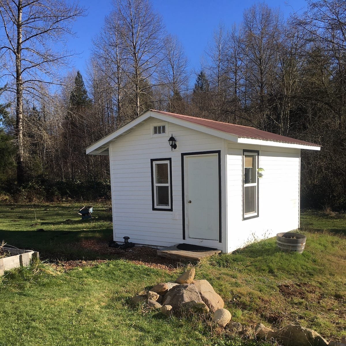 53aa35c3-24e1-48f2-9d0d-1197cf23b9f4 Small white garden shed with a red metal roof, surrounded by green grass and trees in a sunny outdoor setting. The shed features a front door, windows, and a stone pathway leading to it.