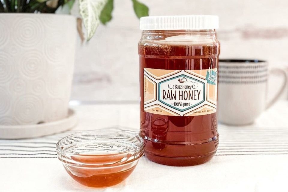 Frey+Family+Farm,+WA+-+all+a+buzz+honey Jar of raw honey labeled "100% pure" next to a small glass bowl filled with honey, set on a striped tablecloth with a potted plant and a mug in the background.
