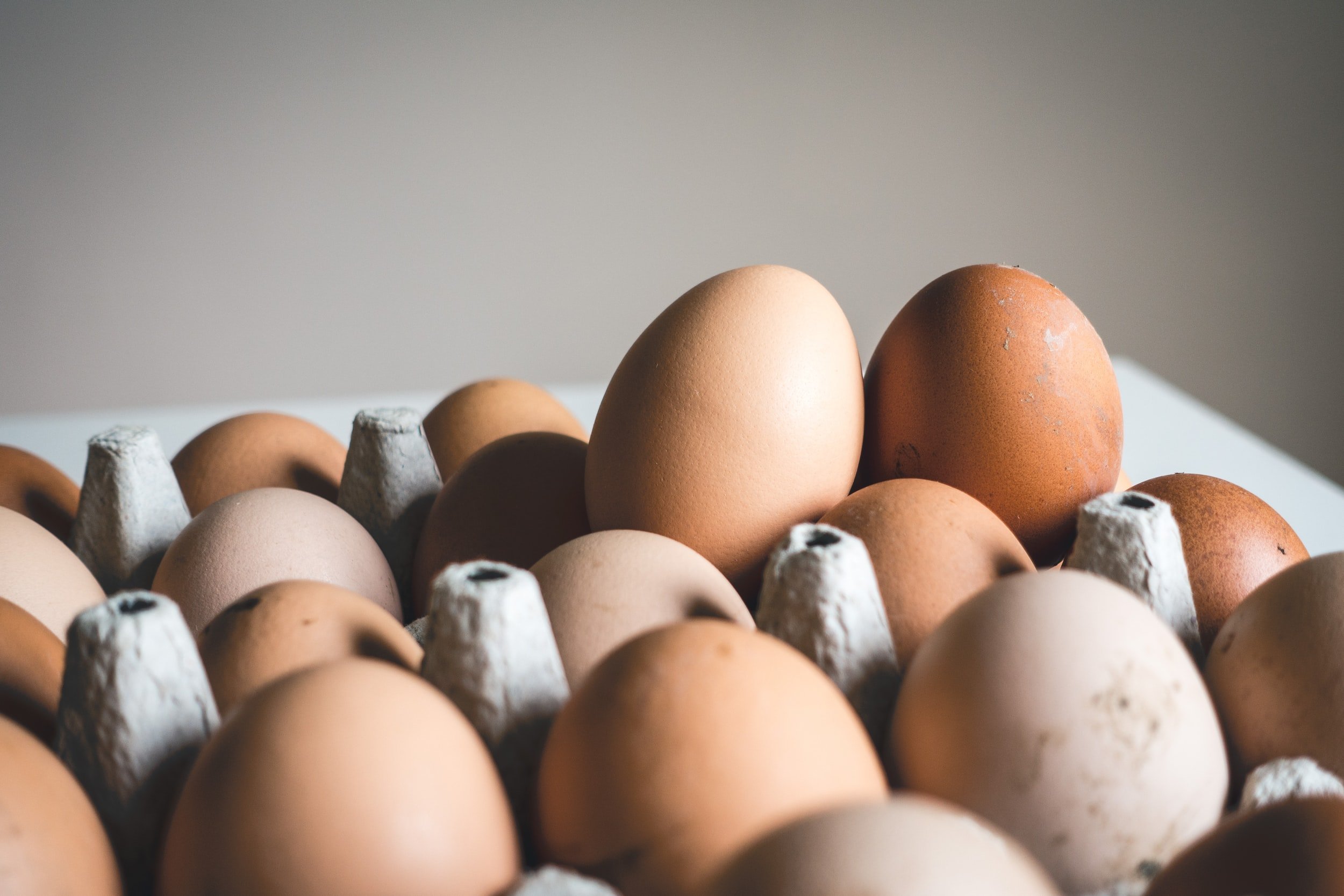 unsplash-image-Hj53USePB1E A close-up view of a carton filled with various eggs, showcasing a mix of brown and light-colored eggs arranged neatly in their compartments. The background features a soft, neutral tone, enhancing the focus on the eggs.